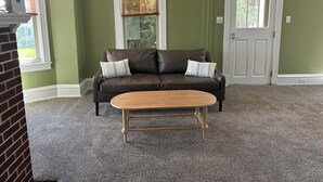 Ein Wohnzimmer mit einer braunen Ledersofa, zwei Kissen, einem Holztisch, grünen Wänden, einer weißen Tür und einem Fenster mit Jalousien.