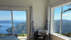 Un dormitorio con cama, una silla y amplias ventanas que ofrecen vistas a un lago y colinas.