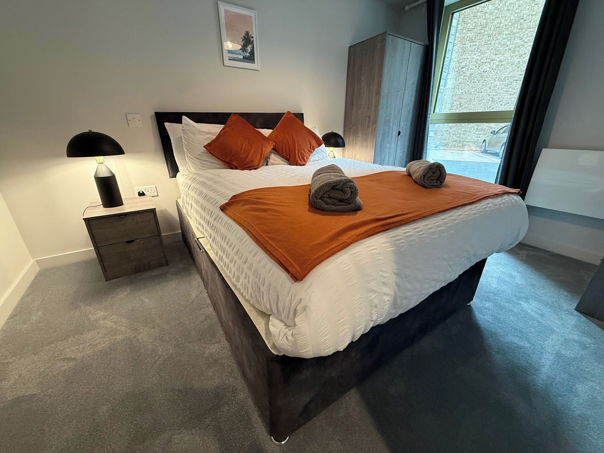 A neatly made bed with a brown blanket and rolled towels, a bedside lamp, a small wooden nightstand, and a large window with a view of the outside.