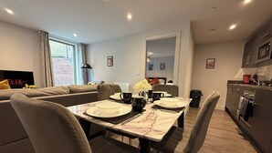 A modern living room with a dining area, a sofa, a TV, and a kitchen in the background. - Stylish 2BR Flat •Parking• 6 min to Luton Airport (Luton)
