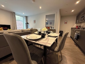 A modern living room with a dining area, a sofa, a TV, and a kitchen in the background. - Stylish 2BR Flat •Parking• 6 min to Luton Airport (Luton)