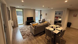 A modern living room with a sofa, a TV, a dining table with chairs, and a large window. - Stylish 2BR Flat •Parking• 6 min to Luton Airport (Luton)