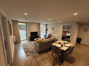 A modern living room with a sofa, a TV, a dining table with chairs, and a large window. - Stylish 2BR Flat •Parking• 6 min to Luton Airport (Luton)