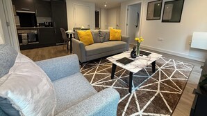 A modern living room with a grey sofa, a coffee table with a vase of flowers, and a kitchen in the background. - Stylish 2BR Flat •Parking• 6 min to Luton Airport (Luton)