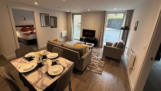 A modern living room with a dining area, a sofa set, a TV, and a balcony with a view. - Stylish 2BR Flat •Parking• 6 min to Luton Airport (Luton)