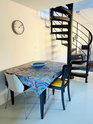 Ein Essbereich mit Tisch, bedeckt mit einer gemusterten Tischdecke, zwei Stühlen, einer blauen Schüssel und einer Wanduhr.