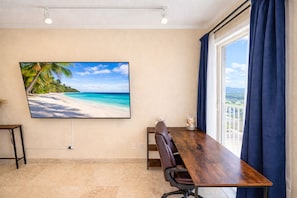 Una sala conferenze con una grande TV a schermo piatto che mostra una spiaggia, un tavolo di legno, una poltrona in pelle marrone e vista sull'esterno attraverso ampie finestre.