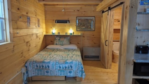 A wooden cabin room with a bed, a small heater, a toilet, and a kitchenette. - "Seacoast" Cabin at Keyser Pond Campground (Henniker)
