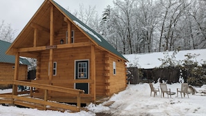 A wooden cabin with a porch, a covered outdoor seating area, and a snow-covered landscape. - "Seacoast" Cabin at Keyser Pond Campground (Henniker)