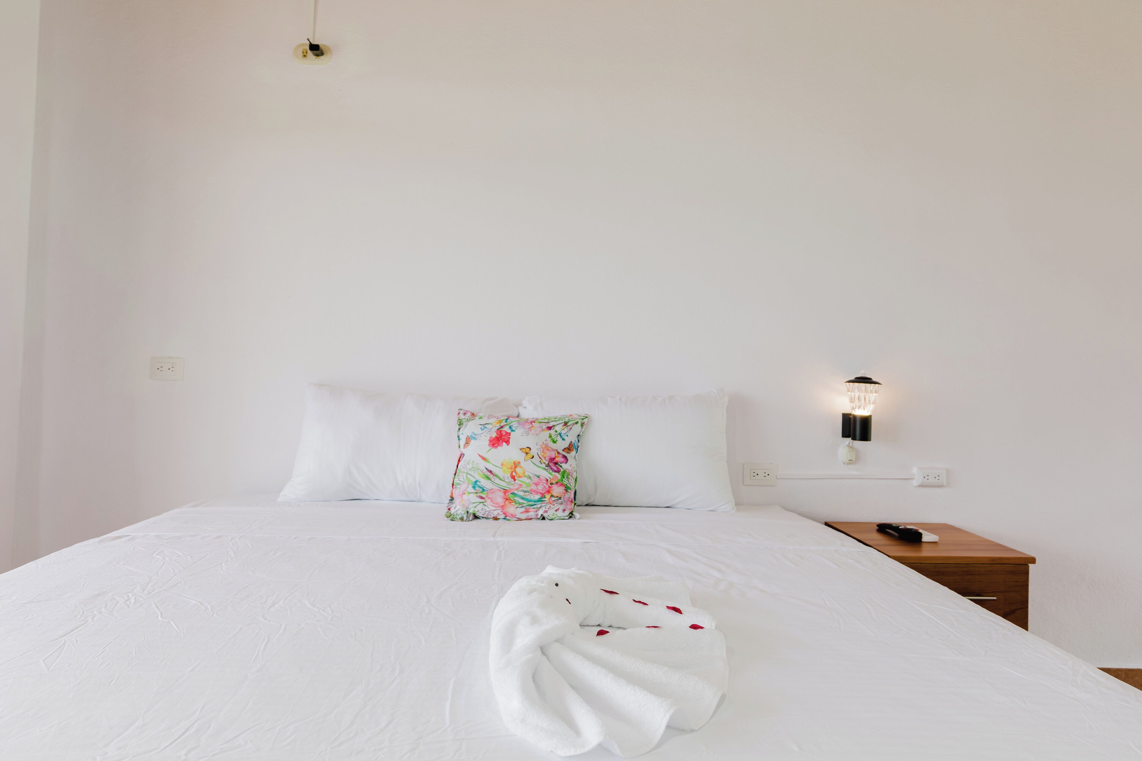 A neatly made bed with a white duvet, a floral pillow, and a towel on the bed. There is a bedside table with a remote control and a wall-mounted light fixture.
