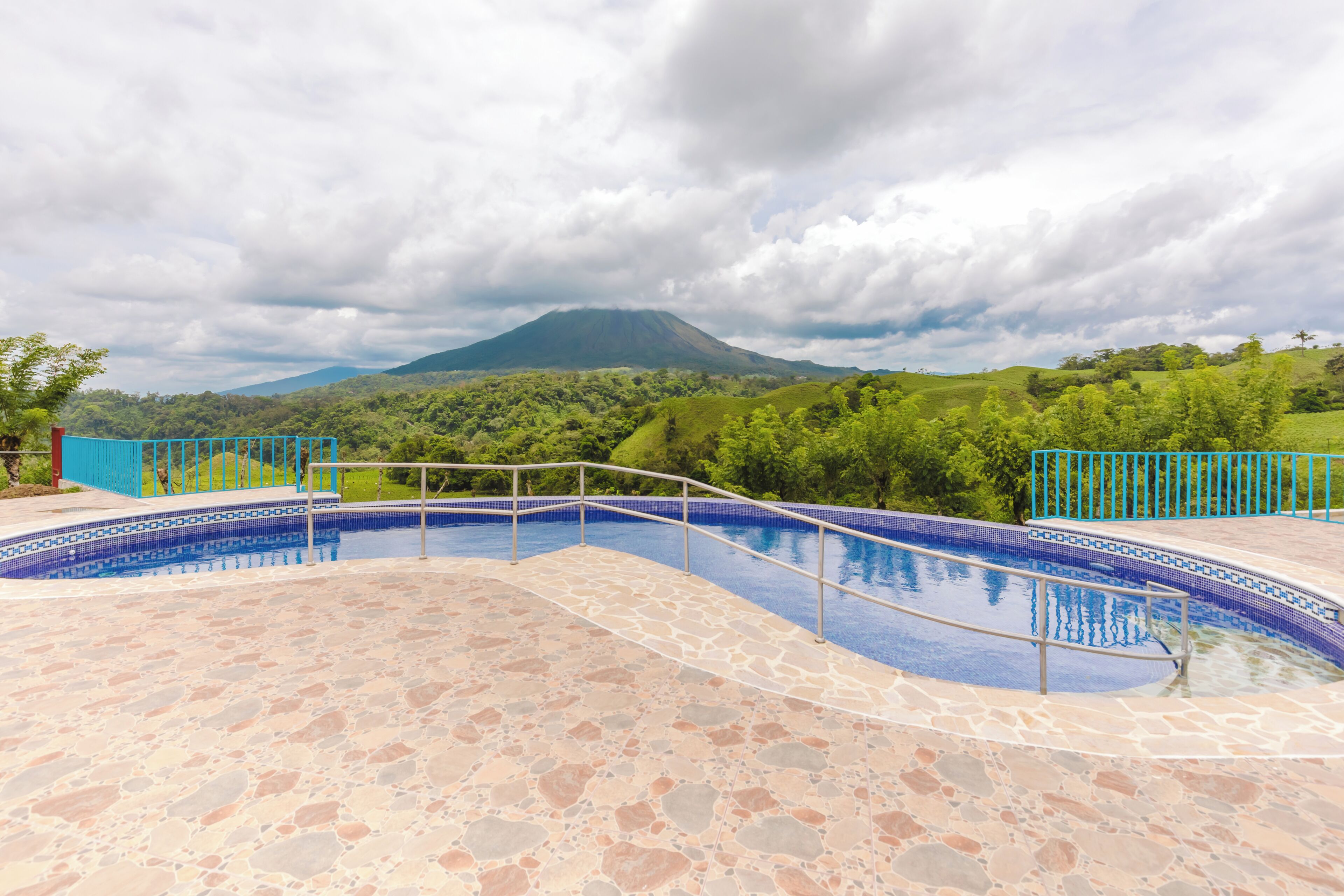 Une piscine entourée d’une terrasse dallée de pierres, délimitée par une clôture bleue, et offrant une vue sur un paysage verdoyant avec une montagne à l’horizon.