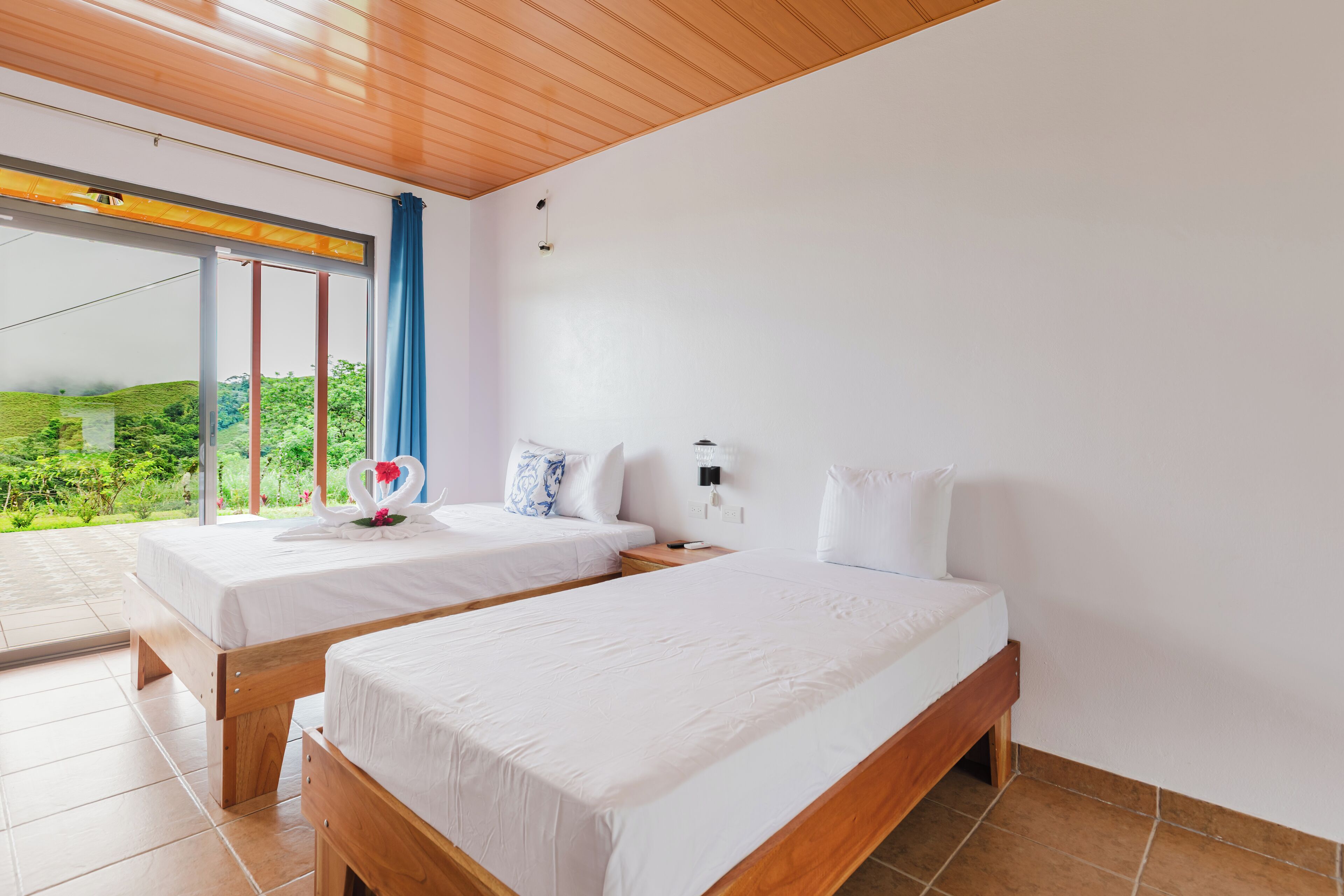 A bedroom with two beds, a large window with a view of greenery, and a wooden ceiling.