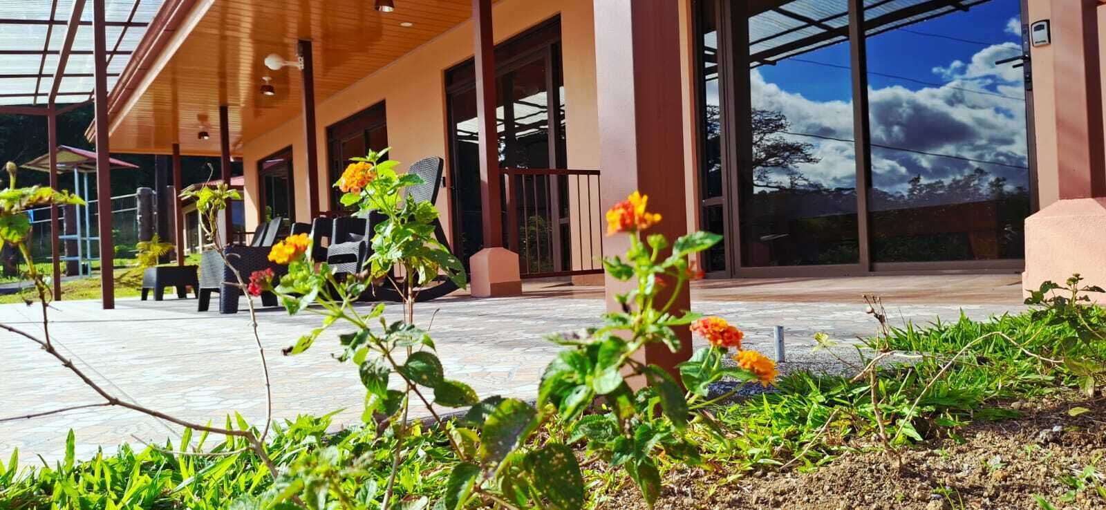 Un edificio con amplios ventanales, un espacio al aire libre cubierto con sillas y un jardín con flores.