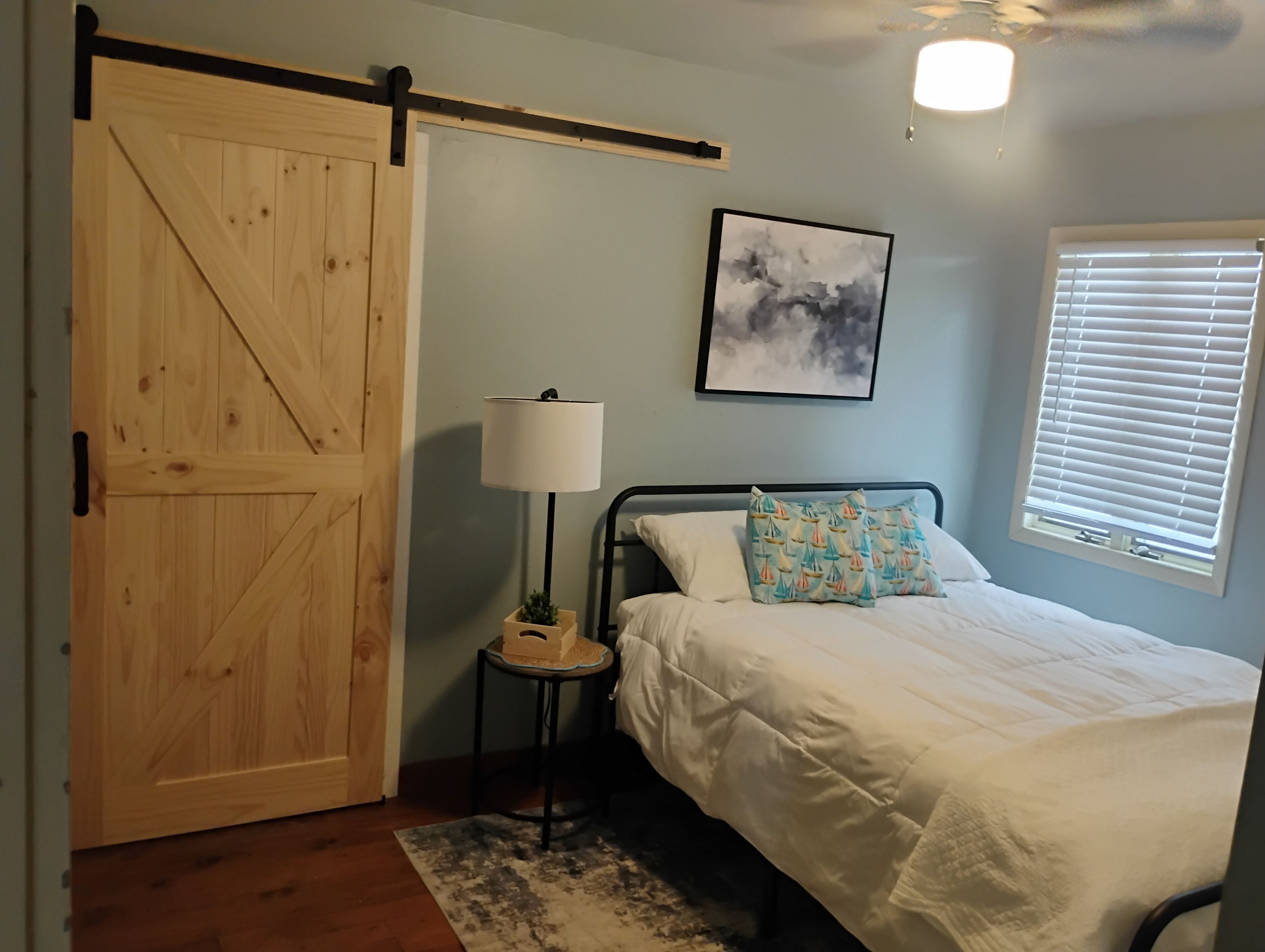 A bedroom with a bed, a nightstand, a lamp, a wooden door, a ceiling fan, and a window with blinds.