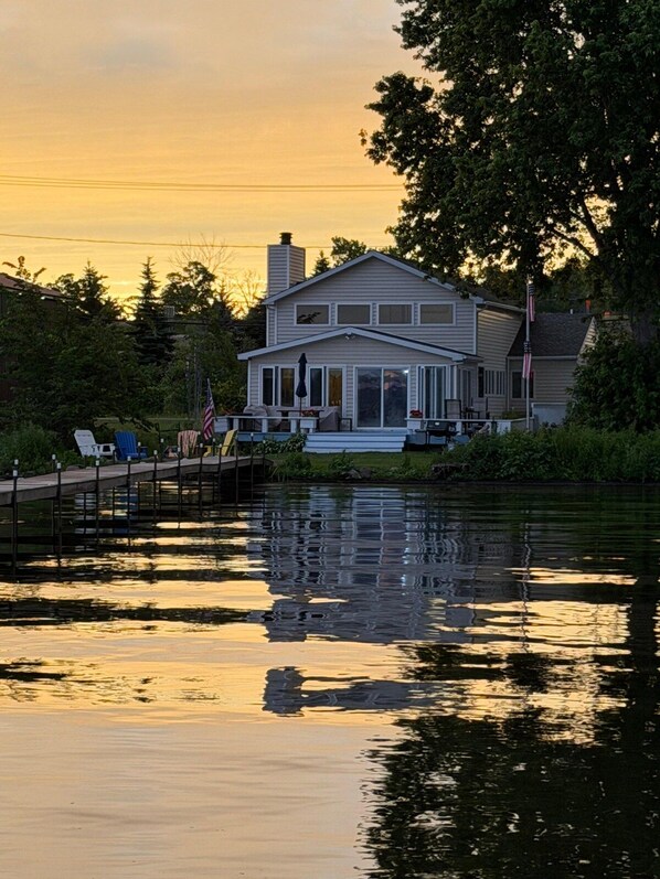A two-story house with a dock extending into a calm body of water, surrounded by greenery.
