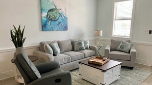 Ein Wohnzimmer mit grauem Sofa, zwei Sesseln, einem Couchtisch und einem Wandbild einer Schildkröte.
