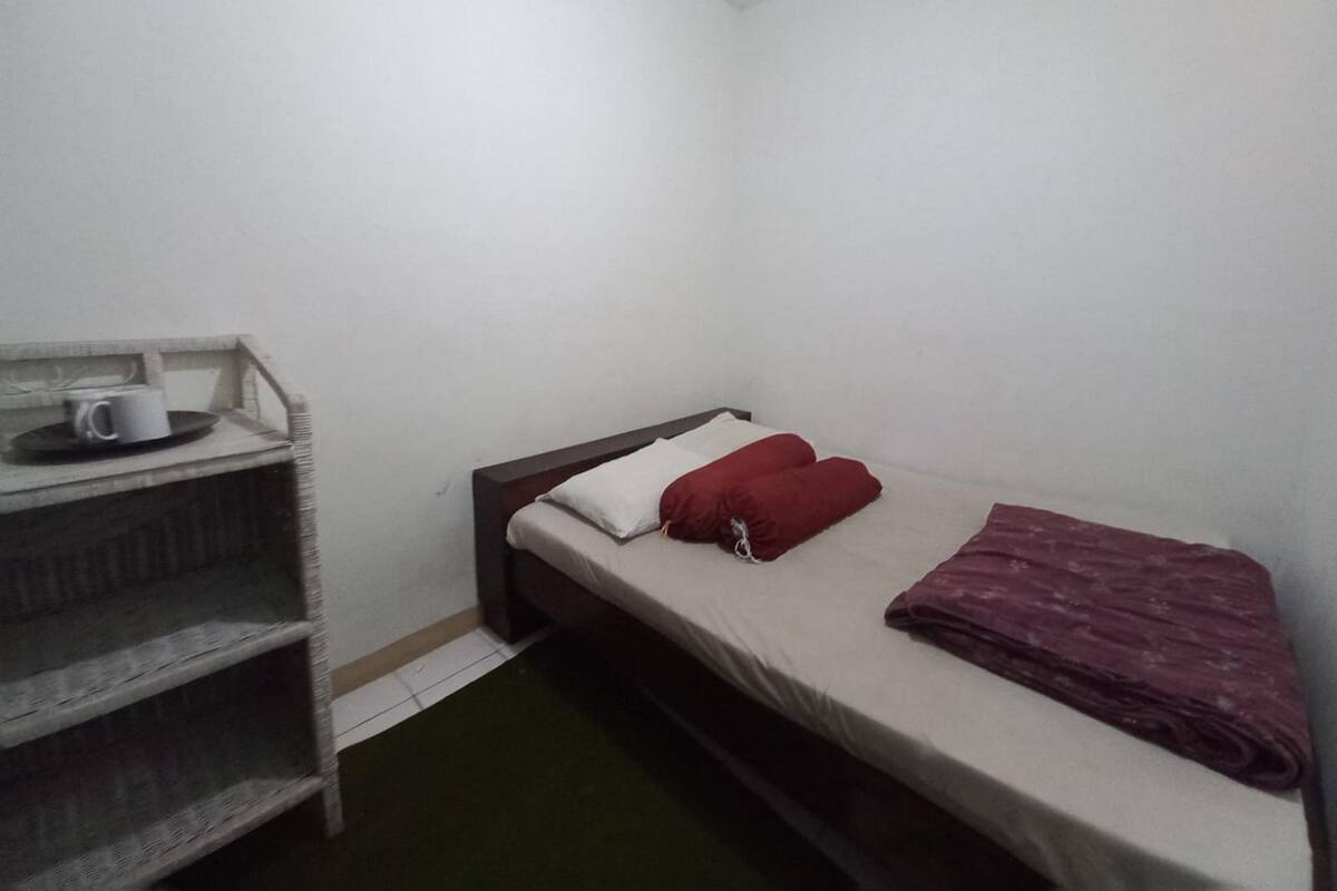 A small, single-bed room with a wooden headboard, a white mattress, and a red pillow. There is a small shelf with a white mug and a plate. The room has white walls and a green floor.