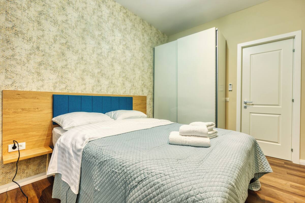 A neatly made bed with a blue headboard, a wooden bedside table, and a wall-mounted power outlet.