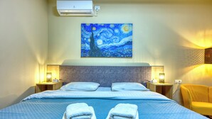 Una camera d'albergo con un letto, due comodini, un condizionatore d'aria a parete, un quadro e una poltrona gialla.
