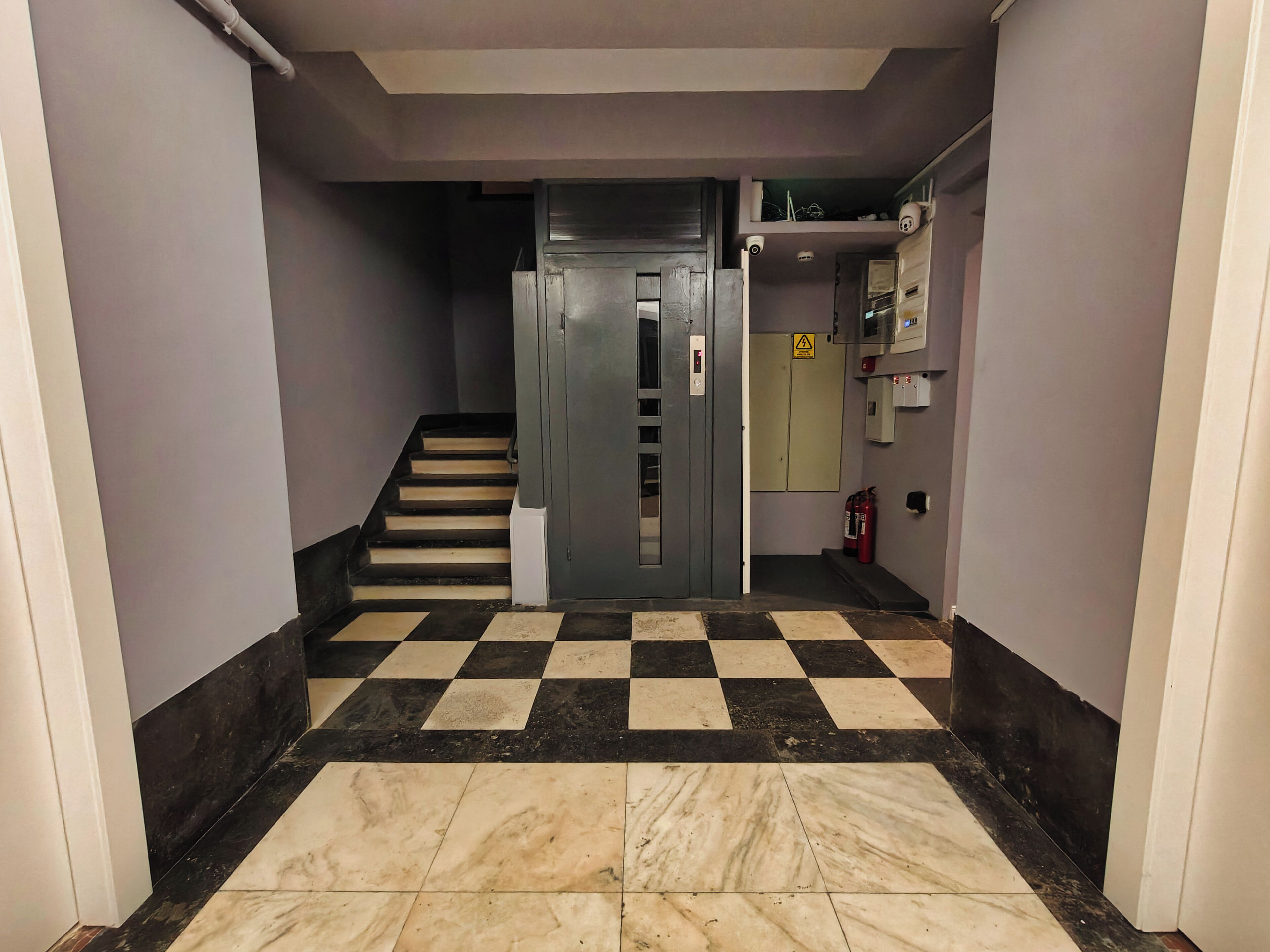 Un couloir avec un sol à carreaux, un ascenseur gris et un extincteur fixé au mur.
