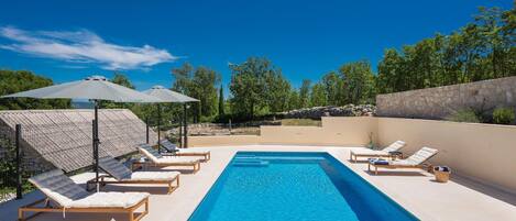 Uma piscina retangular com espreguiçadeiras e guarda-sóis, rodeada por um muro de pedra e árvores.