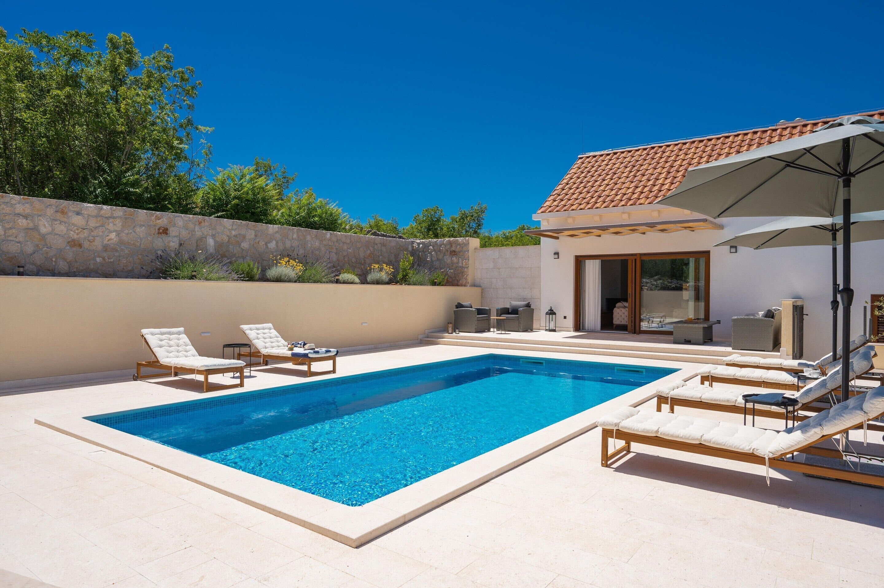 Ein rechteckiger Pool mit Liegestühlen, einer Steinmauer und einem Gebäude mit Ziegeldach.