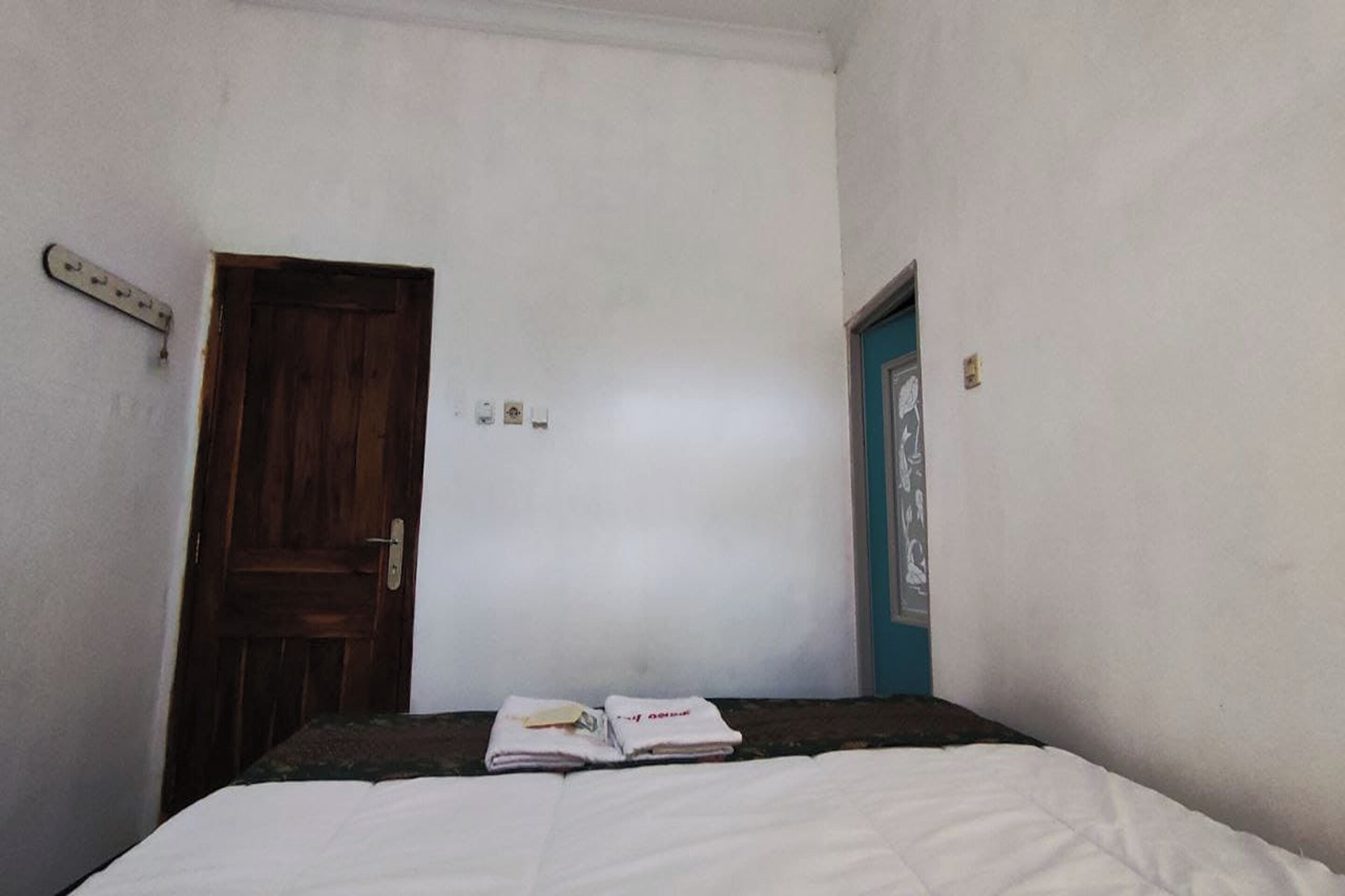 Ein kleines, schlichtes Zimmer mit einem Einzelbett, einer Holztür und einem an der Wand befestigten Lichtschalter.
