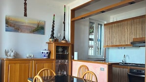 Een eetgedeelte met een glazen tafel en houten stoelen, een keuken met houten kastjes, en een schilderij van een strandgezicht aan de muur.