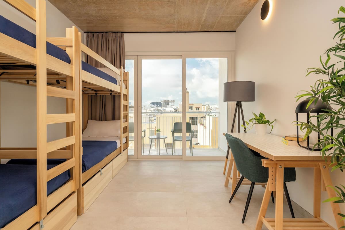 Une chambre avec des lits superposés, un bureau, une chaise, une lampe et une vue sur la ville grâce à de grandes fenêtres.