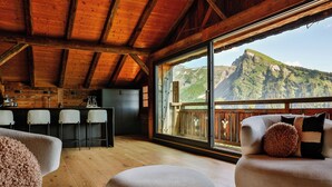 Ambiente interno in legno con cucina, angolo bar e posti a sedere vicino a una grande finestra con vista sulle montagne.