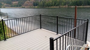 A wooden deck with a metal railing overlooking a calm lake, surrounded by forested hills. - Rustic cabin built 1880s on the Clark Fork River (Clark Fork)