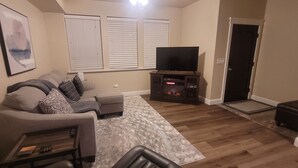 Un salón con sofá, una mesa de centro, un televisor de pantalla plana sobre un mueble, chimenea y una puerta que da a otra habitación.