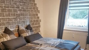 A bedroom with a brick-patterned wall, a bed with grey bedding, a white rug, and a window with curtains.