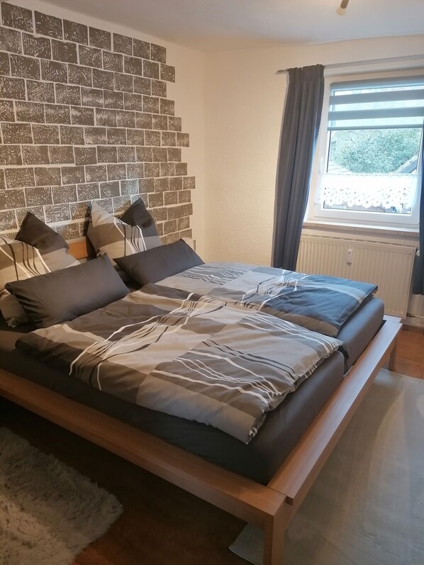 Un dormitorio con pared de ladrillo, cama con ropa de cama gris, alfombra blanca y ventana con cortinas.