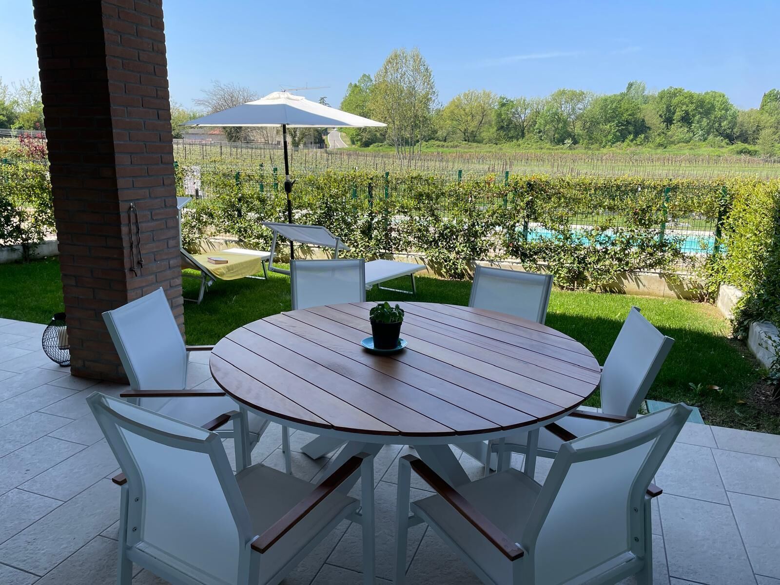 A patio with a round table and white chairs, a brick pillar, and a view of a vineyard and pool in the background.