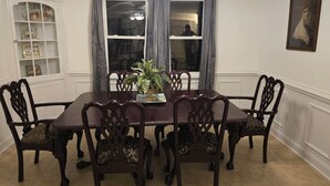 A dining room with a large wooden table surrounded by ornate chairs, a potted plant, and a window with curtains. -  Beautiful 3-bedroom 3 bath home  in the quaint fishing village of Engelhard. (Engelhard)