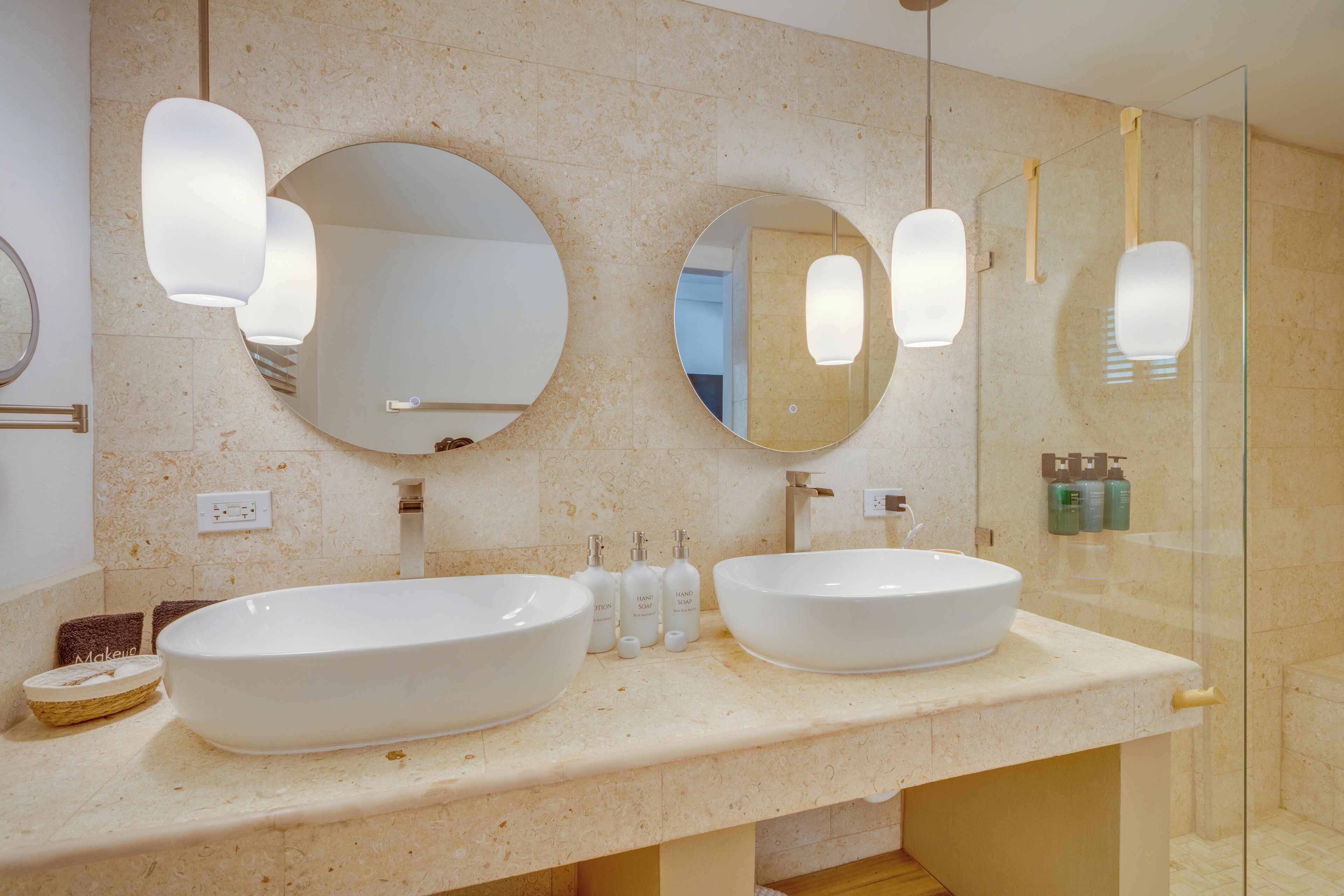 Un baño moderno con dos lavamanos grandes de color blanco, un espejo redondo y una cabina de ducha de vidrio.