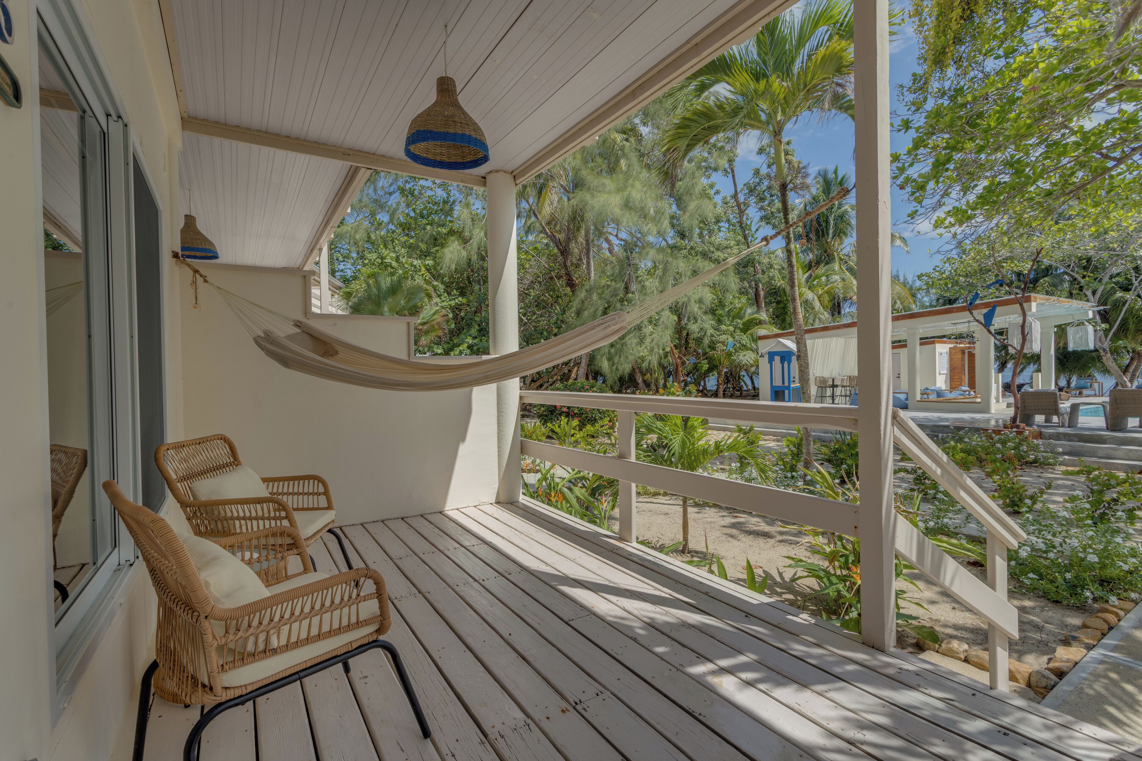 A wooden deck with wicker chairs and a hammock, overlooking a garden and a small building.