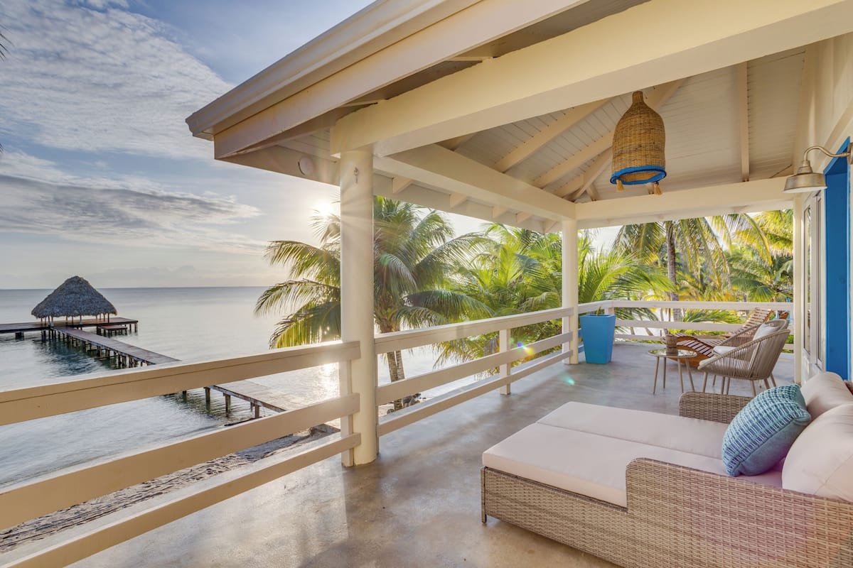 A covered outdoor seating area with a view of the ocean, a thatched hut, and palm trees.