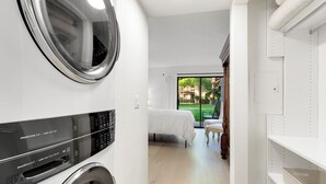 Een wasruimte met een wasmachine en droger, een bed en uitzicht op een tuin via een glazen deur.