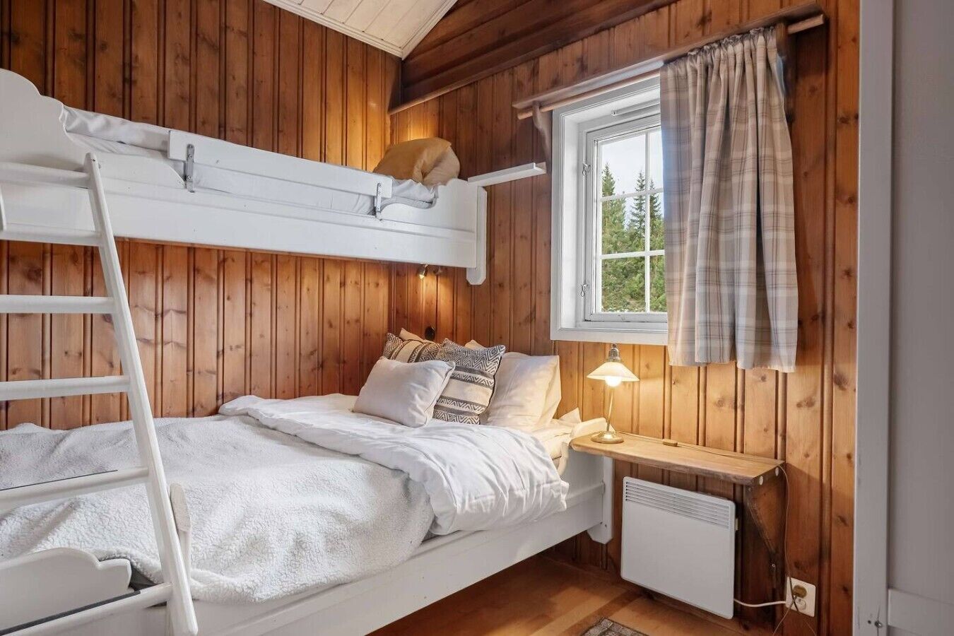 A bunk bed with a ladder, a bedside table with a lamp, a window with curtains, and a radiator.