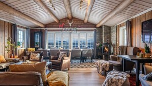 A cozy interior with a wooden ceiling, a fireplace, and a view through large windows. - Trysil Ski-in/out | 6 BR | Sauna (Trysil)