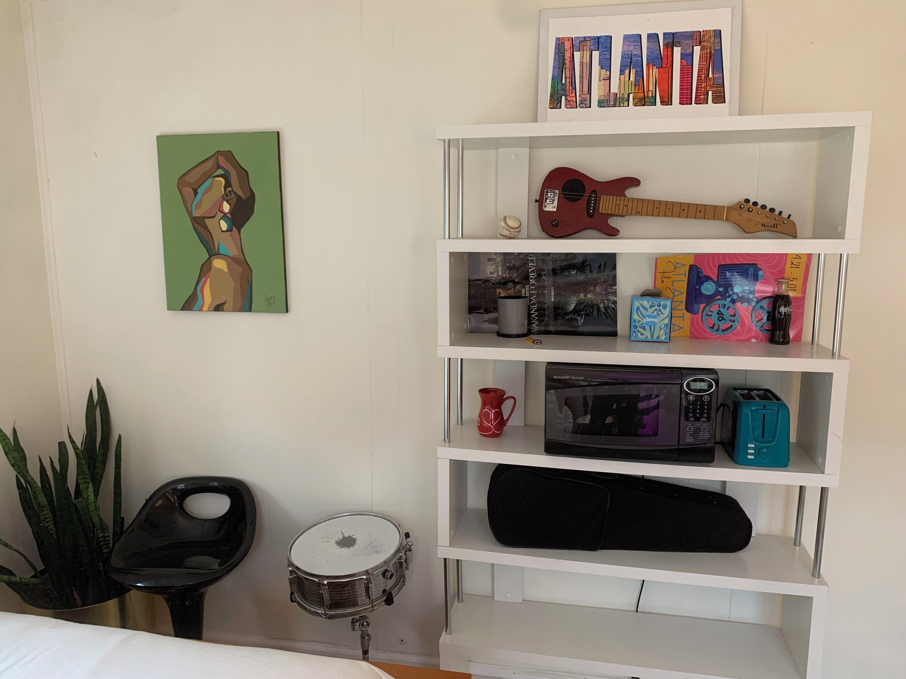 A room with a white wall, a shelf with various items including a guitar, a drum, and a framed picture.