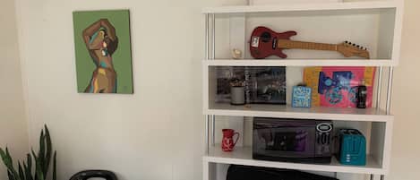 A room with a white wall, a shelf with various items including a guitar, a drum, and a framed picture.