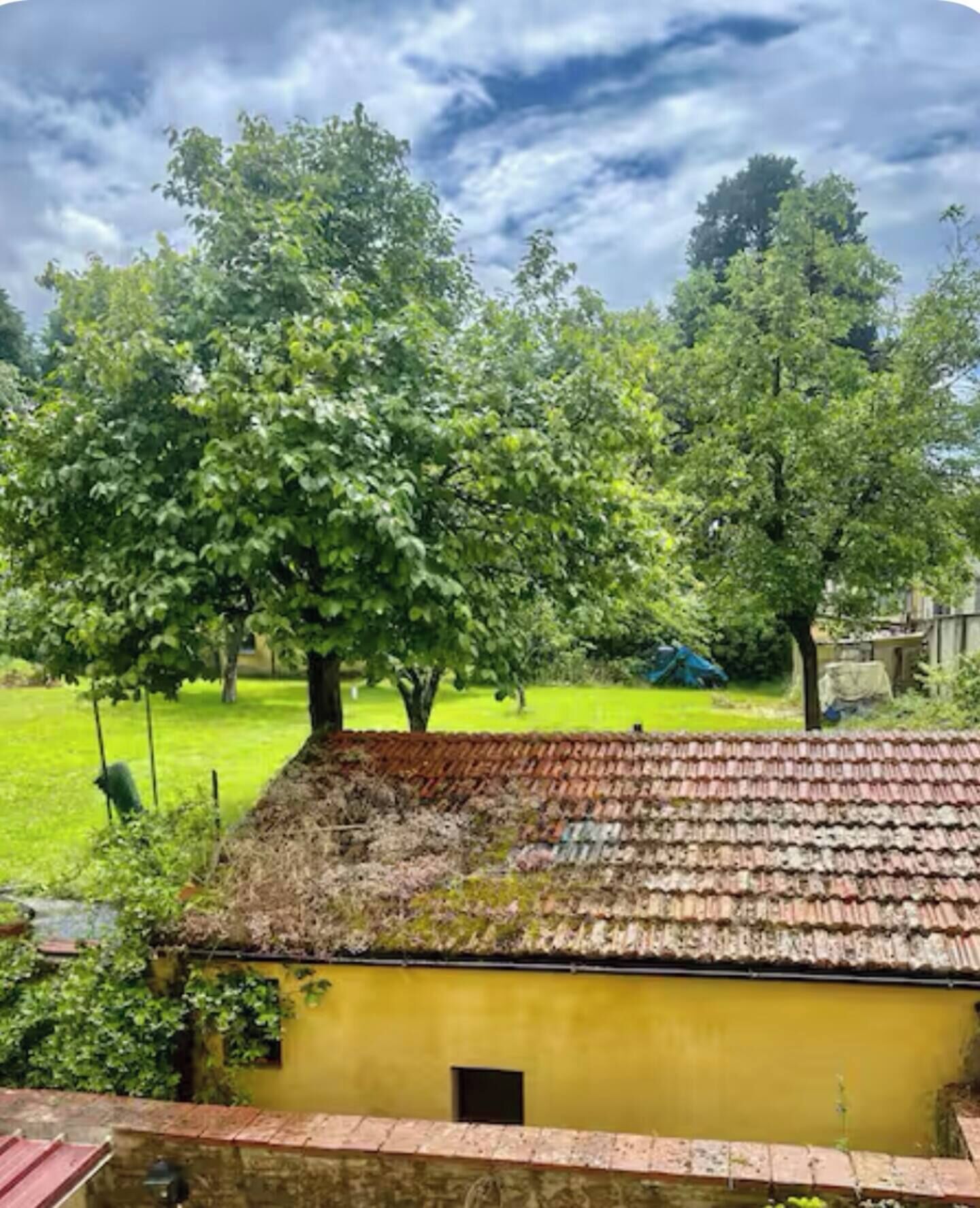 Un bâtiment rustique à la façade jaune et au toit de tuiles, entouré d’arbres verdoyants et d’une cour gazonnée.