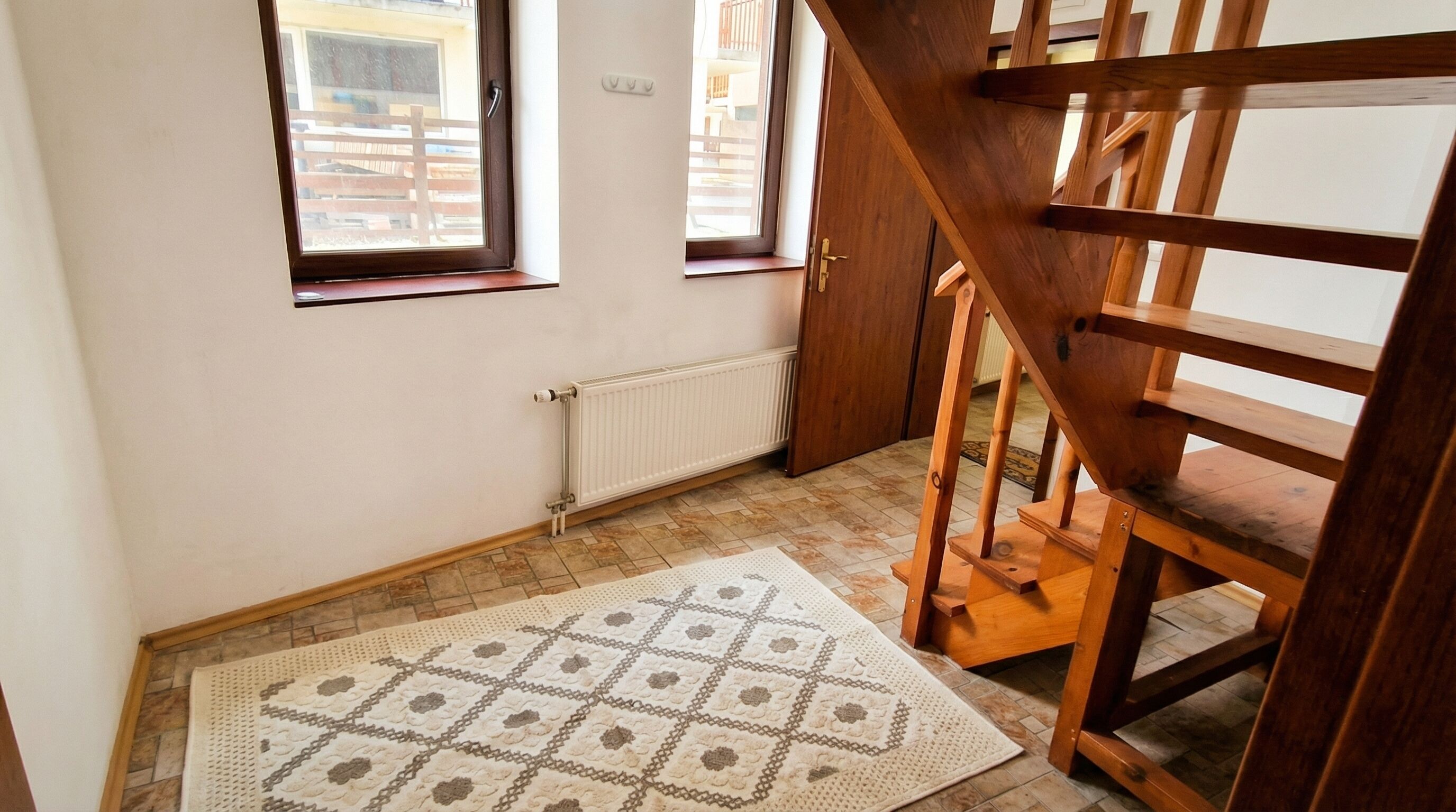A room with a wooden staircase, two windows, a radiator, and a patterned rug.