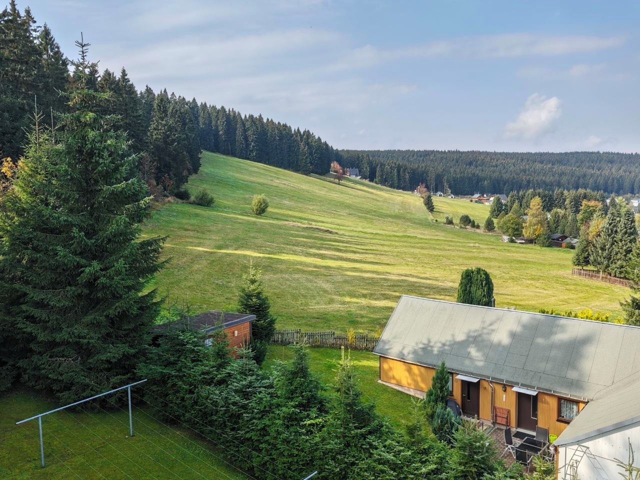 Een landelijk landschap met een huis, een omheind erf en een bebost heuvel op de achtergrond.