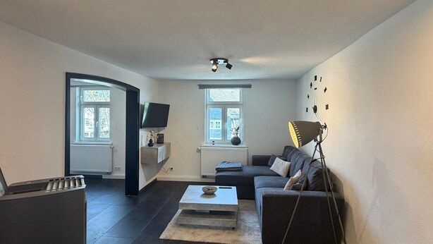 A modern living room with a dark sofa, a coffee table, and a TV. There is a window with blinds, a lamp, and a ceiling light. - My Altstadt Domizil - Domizil "Freiraum" (Hallenberg)