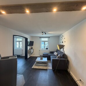 Un salon moderne avec un canapé foncé, une table basse et un téléviseur. On y trouve une fenêtre avec des stores, une lampe et un luminaire au plafond.