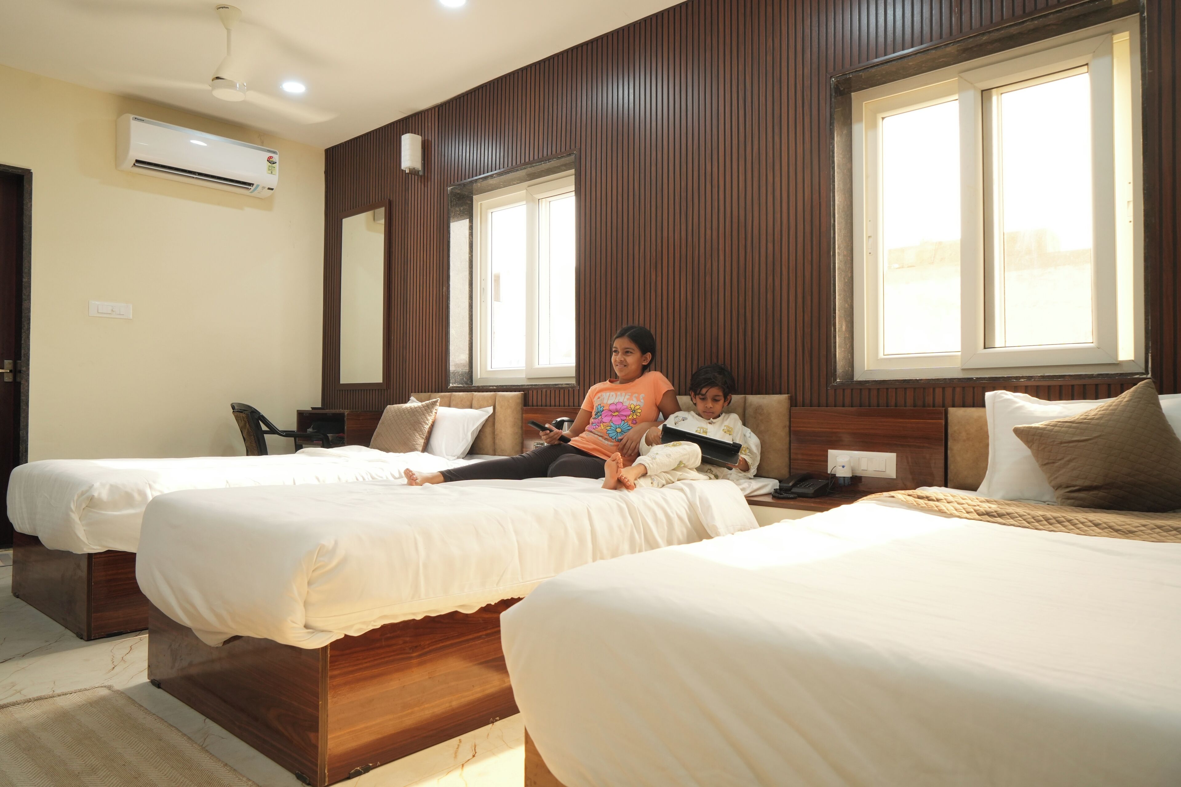 Habitación de hotel con dos camas individuales, paneles de madera, ventanas amplias, un equipo de aire acondicionado y una mesita de noche con lámpara.